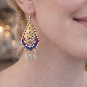 May include: A teardrop-shaped earring with a floral design. The earring features a copper-colored border, a gold center, and a blue and red floral pattern. Three silver-colored dangles hang from the bottom. The earring is worn on a person's ear.