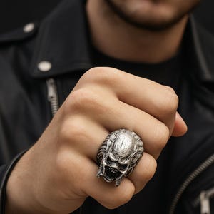 Puede incluir: Un anillo plateado con un diseño detallado de calavera. El anillo tiene una superficie texturizada y se lleva en un dedo. El fondo es oscuro y la persona lleva una chaqueta de cuero negra.