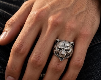 Könnte beinhalten: Ein silberfarbener Metallring mit einem detaillierten, dreidimensionalen Bärenkopf-Design. Der Bär ist mit einer strukturierten Felloberfläche, spitzen Ohren und einem geöffneten Maul dargestellt, was die handwerkliche Präzision zeigt.