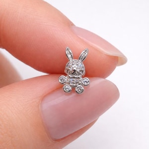 May include: A miniature, silver-toned rabbit charm, embellished with sparkling gems. The bunny features long ears, a rounded body, and small, circular paws. The charm is held between fingers, highlighting its detailed design and craftsmanship.
