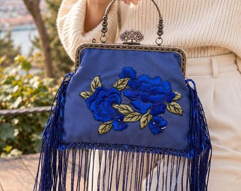 Può includere: Una borsa a mano in raso blu con intricati ricami floreali blu scuro e foglie dorate. La borsa presenta una cornice metallica con una chiusura decorativa, un manico curvo e lunghe frange blu scintillanti che pendono dal fondo e dai lati.