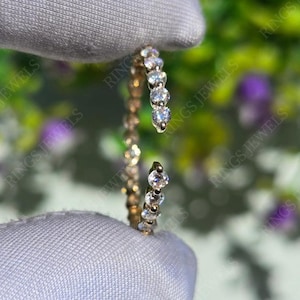Op de afbeelding: Een goudkleurige open ring met een rij kleine, heldere edelstenen. De ring wordt vastgehouden tegen een wazige achtergrond van groen en paars gebladerte. De woorden "RINGS JEWELS" zijn zichtbaar op de achtergrond.
