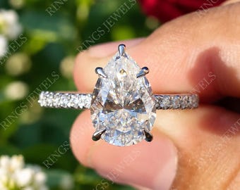 May include: A silver-toned ring featuring a large pear-cut clear gemstone held by six prongs, set on a slender band encrusted with small round clear stones along the sides, showcasing a brilliant faceted surface and a polished metallic finish.