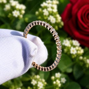 May include: A rose gold eternity band featuring a continuous row of small, round, clear, faceted stones set in a shared prong setting. The ring is thin and circular, showcasing a metallic rose gold finish that reflects light across the stones.