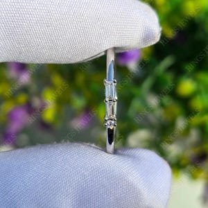 Peut inclure: Une fine bague en argent ornée de trois pierres précieuses : deux rectangulaires et une ronde. Les pierres sont disposées verticalement. La bague est présentée sur un fond blanc et doux. Design minimaliste et élégant.