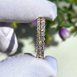 May include: Two gold-colored eternity rings with clear gemstones. The rings are held against a white fabric background, with the words "RINGS JEWELS" visible in the background. The rings are in focus.