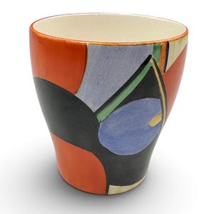 May include: A ceramic cup with a tapered shape and a cream-colored interior. The exterior features a hand-painted abstract design with bold blocks of orange, black, light blue, and green, accented by thin yellow lines and geometric shapes.
