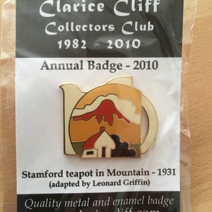May include: A gold-colored enamel pin featuring a teapot with a red mountain scene. The pin is a collector's badge for the Clarice Cliff Collectors Club, 1982-2010. The text on the pin reads "Stamford teapot in Mountain - 1931 (adapted by Leonard Griffin)" and "Quality metal and enamel badge www.claricecliff.com".