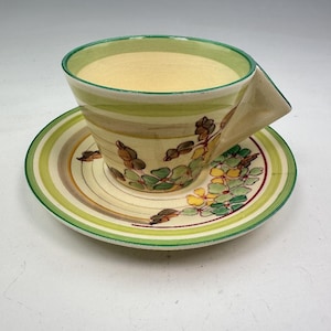 Op de afbeelding: Een vintage theekop en schotel set. De kop heeft een hoekige handgreep en is versierd met groene, gele en bruine bloemmotieven. De schotel heeft bijpassende strepen en bloemaccenten, met een groene rand. De set is crème van kleur.