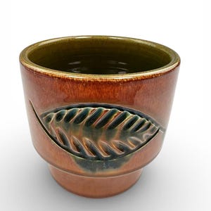 May include: A ceramic planter with a leaf design. The planter is a warm brown color with a green interior and a leaf-shaped cutout on the side. The leaf cutout is filled with a darker green glaze.