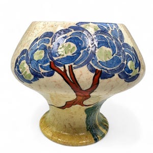 Puede incluir: Un jarrón decorativo de cerámica con una forma y diseño únicos. El jarrón presenta una base de color crema con diseños florales azules y verdes. Un diseño de árbol rojo está pintado en la parte delantera. El jarrón tiene una parte superior ancha y una base de pedestal.