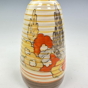 Puede incluir: Un salero de cerámica con forma ovalada y redondeada. El salero presenta una base de color crema con rayas horizontales amarillas y naranjas. El diseño incluye motivos florales estilizados en naranja, amarillo y gris. La parte superior tiene varios agujeros pequeños.