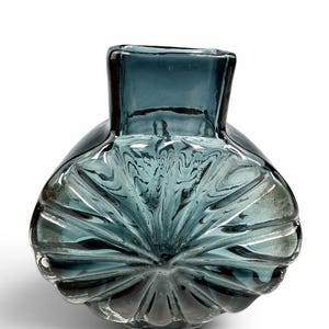 Könnte beinhalten: Eine teal-farbene Glasvase mit einem einzigartigen, strukturierten Design. Die Vase hat eine quadratische Oberseite und einen abgerundeten, gerippten Boden. Das Innere der Vase weist ein wirbelndes Muster auf. Das Glas scheint transparent zu sein.
