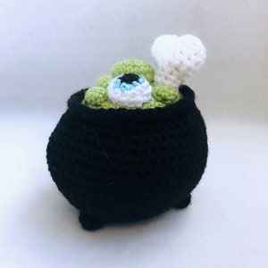May include: A black crocheted cauldron with a green, eye-shaped design and a white bone. The cauldron is filled with green, eye-shaped objects and a white bone. The eye has a blue iris and black pupil. A whimsical Halloween decoration.