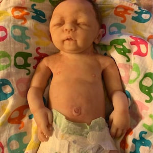 May include: A realistic baby doll with closed eyes, wearing a diaper and lying on a colorful blanket with a pattern of elephants.