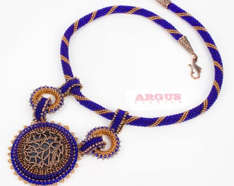 Puede incluir: Un collar de cuentas azules y doradas con un gran colgante redondo que presenta un diseño negro y dorado. El colgante está unido al collar con dos lazos más pequeños de cuentas.