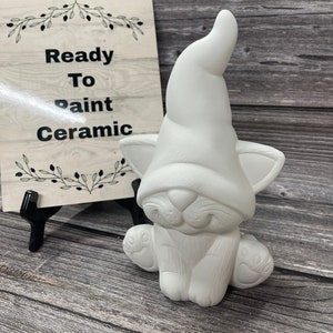 May include: White ceramic cat figurine wearing a Santa hat. The figurine is ready to be painted. The sign in the background reads "Ready To Paint Ceramic".