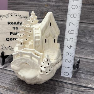 May include: A white ceramic winter village scene, ready to paint. The scene includes a house, bridge, trees, and a water wheel. The piece measures approximately 6 inches tall.