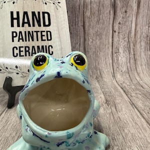 May include: A hand-painted ceramic frog-shaped container in shades of blue, with dark blue and purple accents. The frog has large, yellow-rimmed eyes and an open mouth. A wooden sign behind it reads "HAND PAINTED CERAMIC."