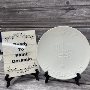 May include: White ceramic plate with the words "Happy Hanukkah" and a menorah design. A sign reads "Ready To Paint Ceramic".