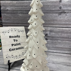 May include: White ceramic Christmas tree with a base and holes for lights. The tree is ready to be painted. The sign says "Ready To Paint Ceramic".