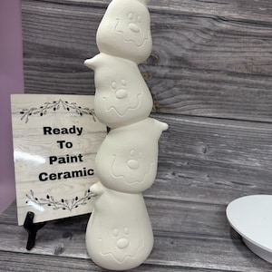 May include: A stack of four unpainted ceramic ghost figures with smiling faces. The ghosts are stacked vertically, creating a whimsical decorative piece. A sign that says "Ready To Paint Ceramic" is in the background.