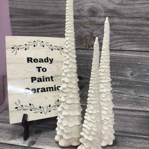 May include: Three unpainted white ceramic cone-shaped trees with textured, layered surfaces. Next to the trees is a rectangular sign with a light wood grain pattern, black leaf illustrations, and the black text Ready To Paint Ceramic.