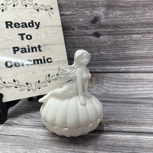 May include: A white ceramic mermaid figurine sitting atop a pumpkin-shaped container. The figurine is unpainted and ready to be painted. A sign in the background reads "Ready To Paint Ceramic".