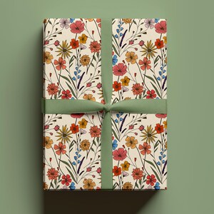 May include: A rectangular gift box wrapped in cream-colored paper featuring a repeating pattern of wildflowers in shades of red, orange, yellow, and blue with green stems, tied with a solid sage green ribbon centered vertically.