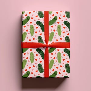 May include: A rectangular gift box wrapped in light pink paper featuring a repeating pattern of green cartoon pickles with smiling faces and small red hearts, tied with a vibrant red ribbon that crosses horizontally and vertically.
