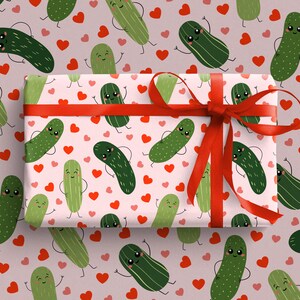 May include: A rectangular gift box wrapped in light pink paper featuring a repeating pattern of smiling green pickles and small red hearts. The box is tied with a vibrant red satin ribbon that is tied into a bow on the right side.