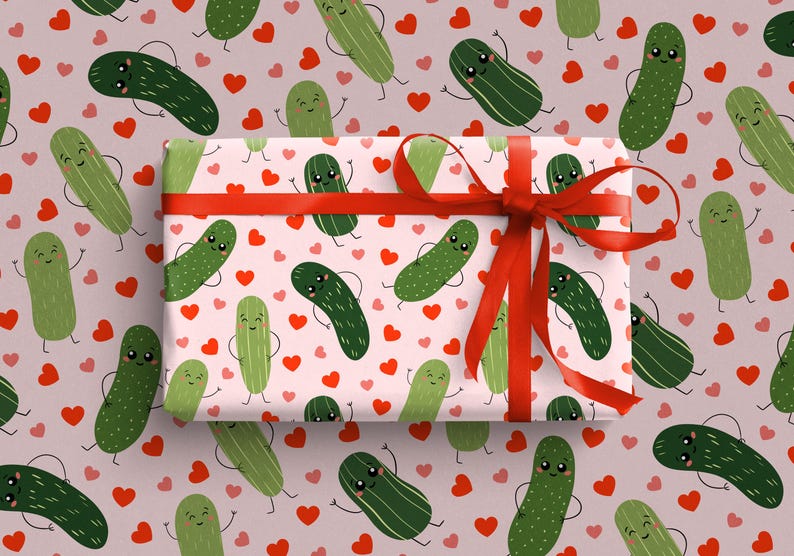 May include: A rectangular gift box wrapped in light pink paper featuring a repeating pattern of smiling green pickles and small red hearts. The box is tied with a vibrant red satin ribbon that is tied into a bow on the right side.