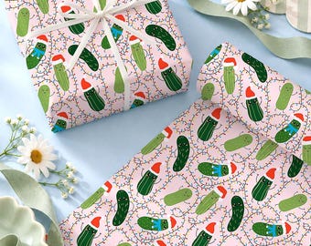 May include: Wrapping paper featuring a repeating pattern of green pickles wearing red Santa hats and blue scarves, interspersed with colorful string lights on a light pink background. A white ribbon is tied around a square gift box.