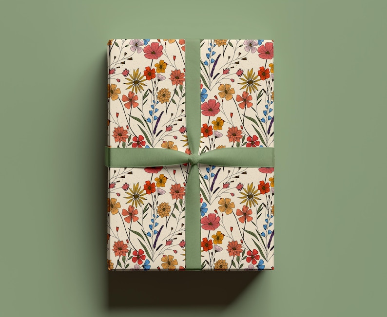 May include: A rectangular gift box wrapped in cream-colored paper featuring a repeating pattern of wildflowers in shades of red, orange, yellow, and blue with green stems, tied with a solid sage green ribbon centered vertically.
