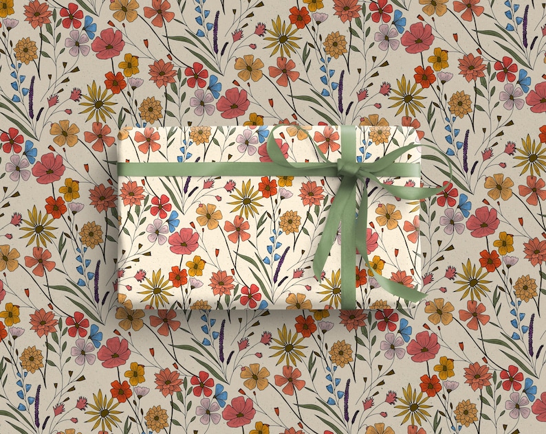May include: A rectangular gift box wrapped in cream-colored paper featuring a repeating pattern of wildflowers in shades of red, orange, yellow, and blue with green stems, tied with a solid olive green ribbon bow centered on top.
