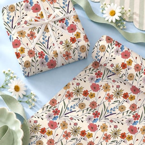 May include: Wrapping paper featuring a repeating pattern of colorful wildflowers in shades of red, orange, yellow, and blue on a cream background. One square gift is wrapped with white ribbon, and a roll of the same paper is unrolled beside it.