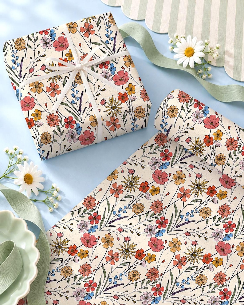 May include: Wrapping paper featuring a repeating pattern of colorful wildflowers in shades of red, orange, yellow, and blue on a cream background. One square gift is wrapped with white ribbon, and a roll of the same paper is unrolled beside it.