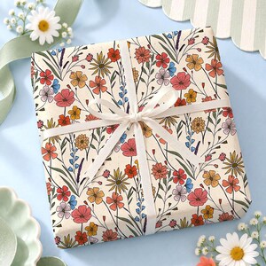 May include: A square gift box wrapped in cream-colored paper featuring a colorful botanical print of red, orange, yellow, and blue wildflowers. The package is tied with a thin white ribbon in a centered bow, creating a clean and simple presentation.