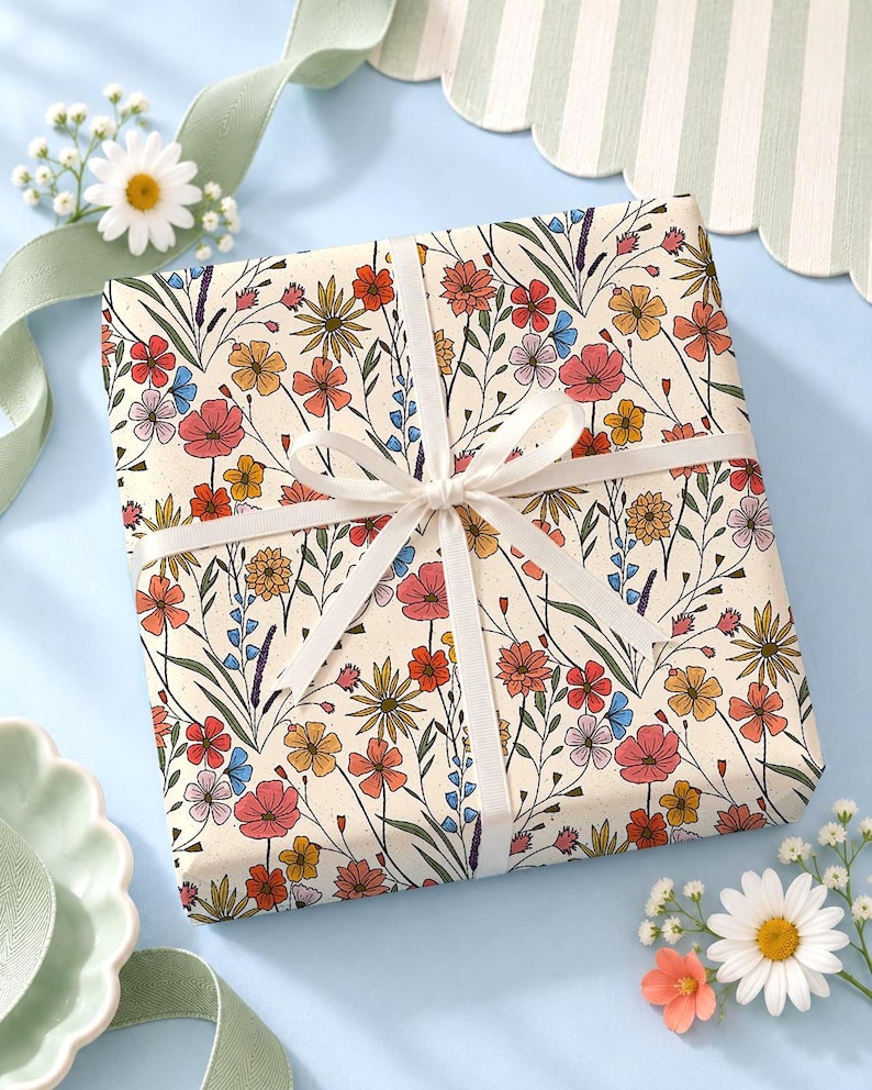 May include: A square gift box wrapped in cream-colored paper featuring a colorful botanical print of red, orange, yellow, and blue wildflowers. The package is tied with a thin white ribbon in a centered bow, creating a clean and simple presentation.