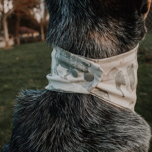 May include: A close-up of a dog wearing a light gray and white bandana with a pattern of stylized animals. The bandana has two snap closures.
