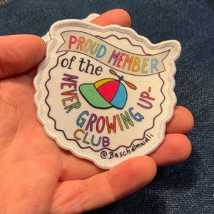 Könnte beinhalten: Weißer gestickter Aufnäher mit einer bunten Cartoon-Illustration eines Hutes mit einem Propeller. Der Text lautet "Proud Member of the Never Growing Up Club" mit dem Text "@BaschAlmaioli" unten.