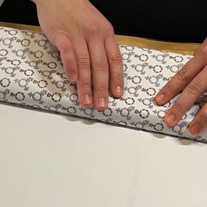 Könnte beinhalten: Eine Person verpackt ein Geschenk mit weißem Geschenkpapier, das ein sich wiederholendes Muster aus violetten und braunen Geschlechtsymbolen aufweist.