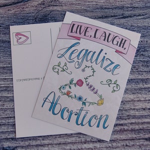 May include: A postcard with a pink banner that says "Live, Laugh, Legalize Abortion". The banner is decorated with flowers and vines. The postcard is white with a pink heart in the top left corner.