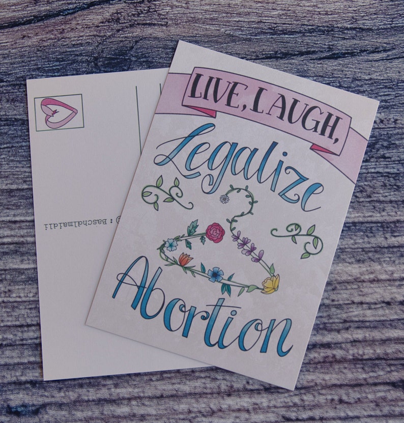 May include: A postcard with a pink banner that says "Live, Laugh, Legalize Abortion". The banner is decorated with flowers and vines. The postcard is white with a pink heart in the top left corner.