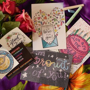 Puede incluir: Cinco tarjetas de felicitaci&oacute;n con dise&ntilde;os coloridos y mensajes escritos a mano. Las tarjetas presentan un coraz&oacute;n con fuegos artificiales, un pastel de cumplea&ntilde;os, un plato con una cuchara, una corona de flores y una pizarra con las palabras "&iexcl;Estoy orgulloso de ti!"