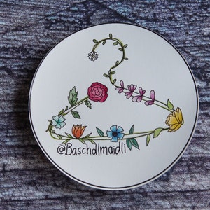 May include: A white ceramic plate with a black outline features a floral design in the shape of a hanger. The flowers are pink, yellow, blue, orange, and white. The text "@Baschdlmaidli" is written in black at the bottom of the plate.