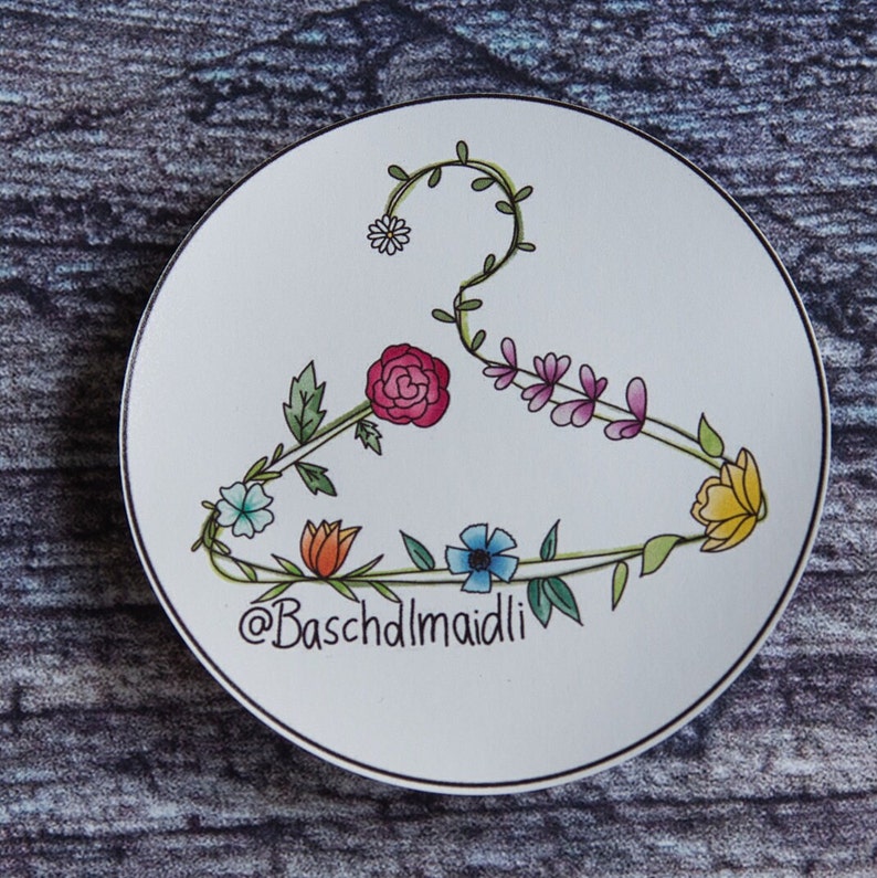 May include: A white ceramic plate with a black outline features a floral design in the shape of a hanger. The flowers are pink, yellow, blue, orange, and white. The text "@Baschdlmaidli" is written in black at the bottom of the plate.