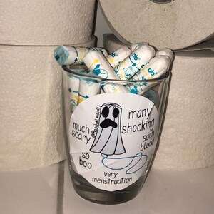 May include: A clear glass filled with tampons, with a sticker featuring a ghost illustration and the words "much scary, so boo, many shocking, such blood, very menstruation". Rolls of toilet paper are in the background.