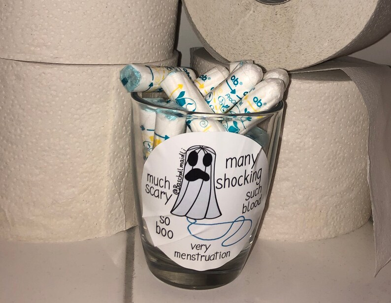 May include: A clear glass filled with tampons, with a sticker featuring a ghost illustration and the words "much scary, so boo, many shocking, such blood, very menstruation". Rolls of toilet paper are in the background.