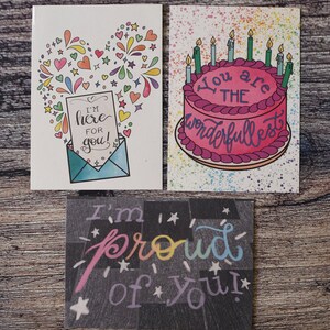 Puede incluir: Tres tarjetas de felicitaci&oacute;n con dise&ntilde;os coloridos. La primera tarjeta tiene una forma de coraz&oacute;n hecha de estrellas y fuegos artificiales coloridos con un sobre y una nota que dice "&iexcl;Estoy aqu&iacute; para ti!" La segunda tarjeta tiene un pastel rosa con velas y el texto "Eres el m&aacute;s maravilloso". La tercera tarjeta tiene un fondo negro con estrellas blancas y el texto "&iexcl;Estoy orgulloso de ti!" en colores arco&iacute;ris.